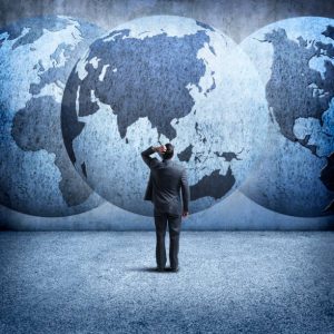 A rear view of a businessman as he stands and looks up at three interlocking globes on the wall in front of him as he tries to sort out the mess of geopolitical events. A globe rotated to Asia is in the center. Map source material courtesy of https://images.nasa.gov/